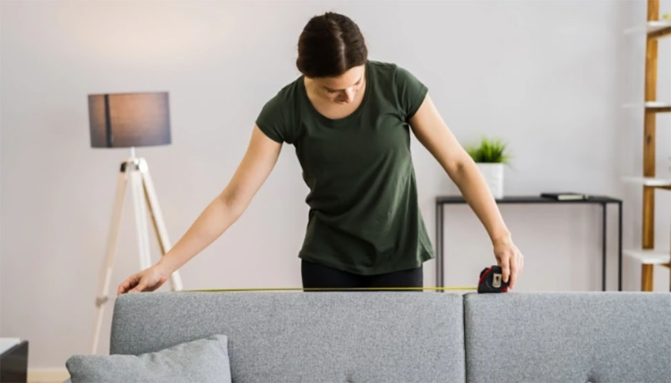 A person measuring a large couch in a cluttered living room, preparing for removal or donation.